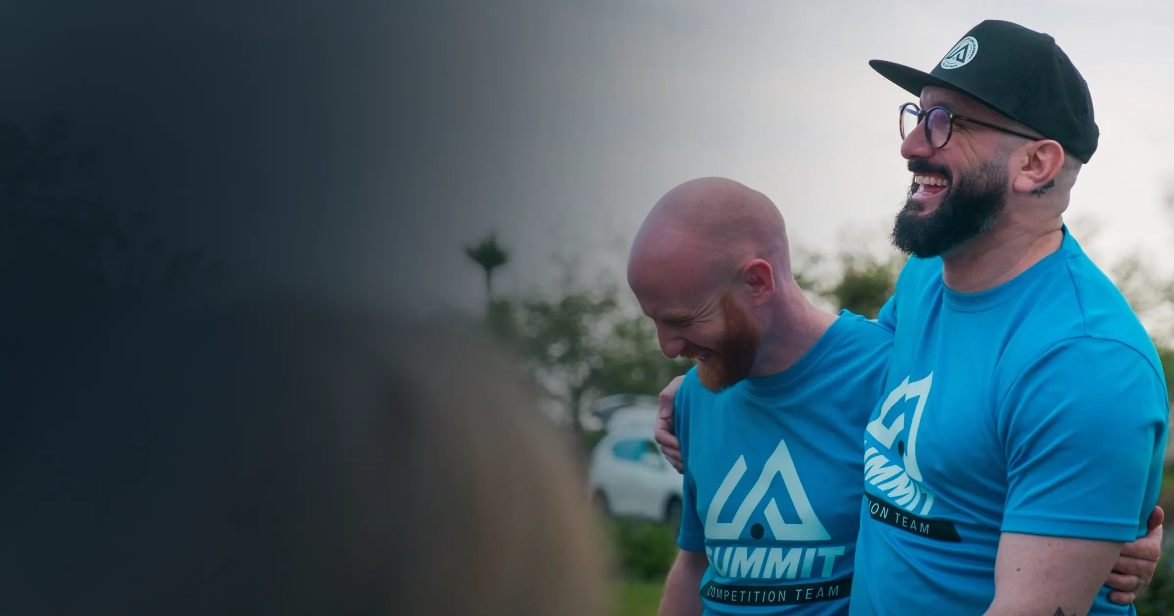 Two smiling men in matching blue t-shirts and caps embrace outdoors, conveying joy and camaraderie.